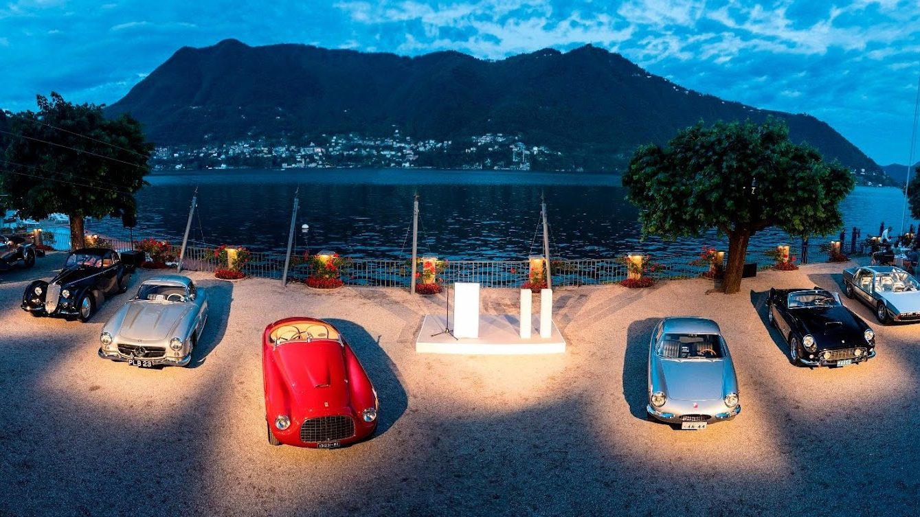 A group of classic cars displayed outdoors near a waterfront, with mountains and a small town in the background under a blue sky—an ideal setting for one of 2025’s top 10 car auctions.