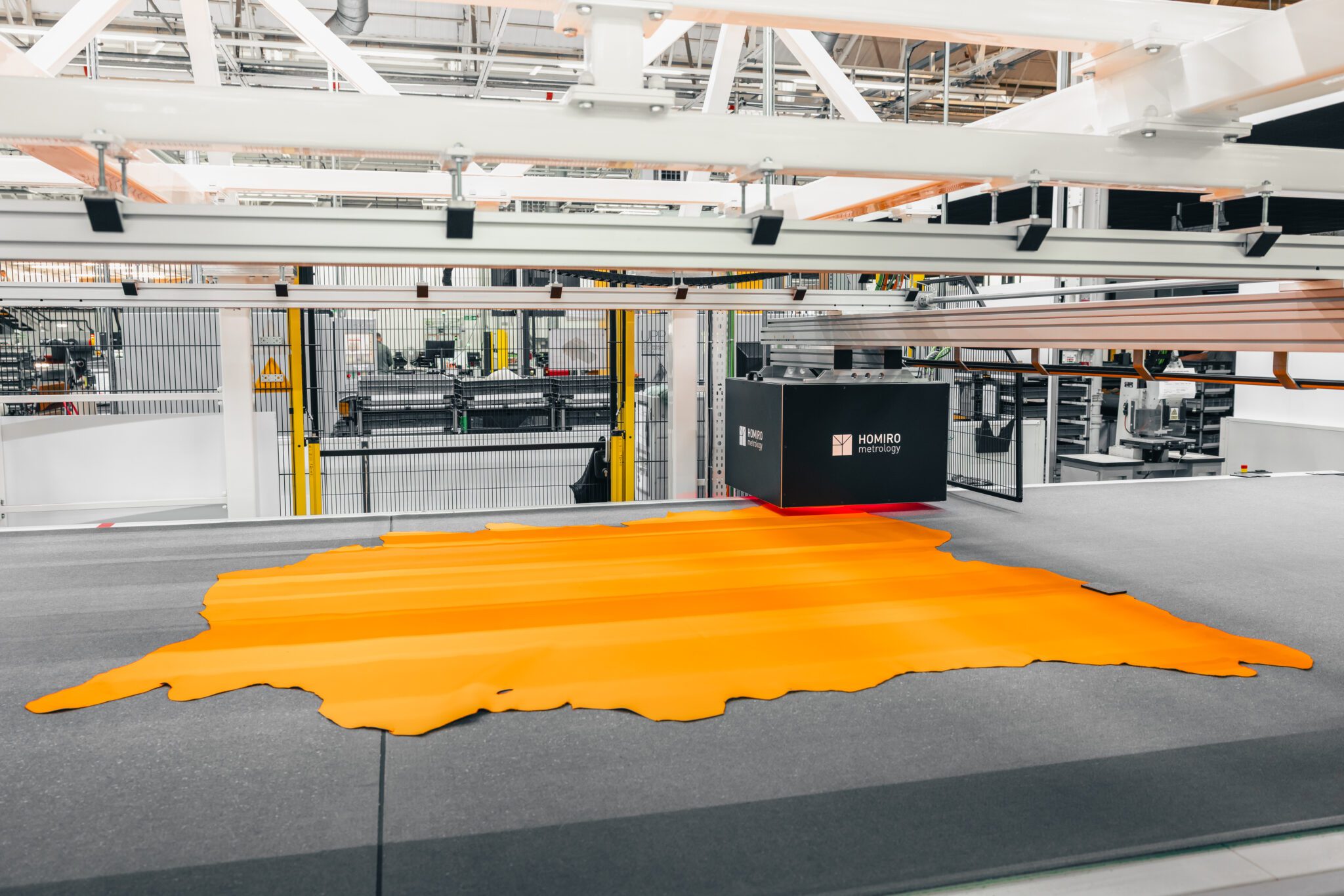 An industrial machine cuts a large piece of orange material in a factory setting, surrounded by metal structures and machinery.