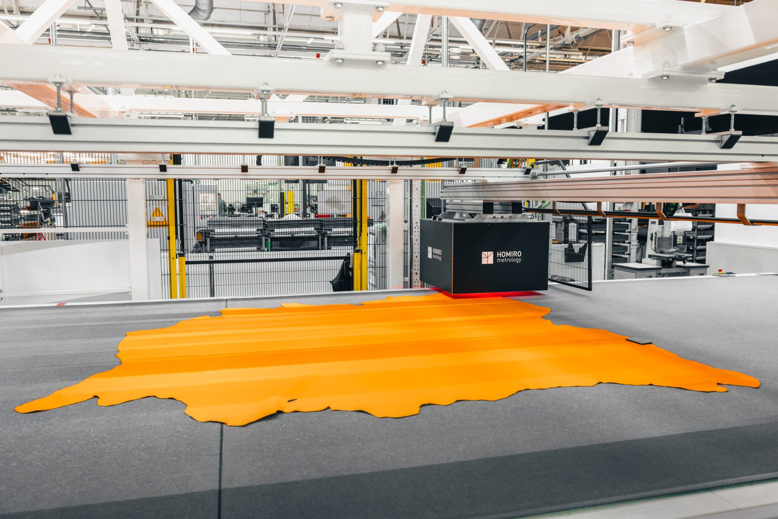 An industrial machine cuts a large piece of orange material in a factory setting, surrounded by metal structures and machinery.