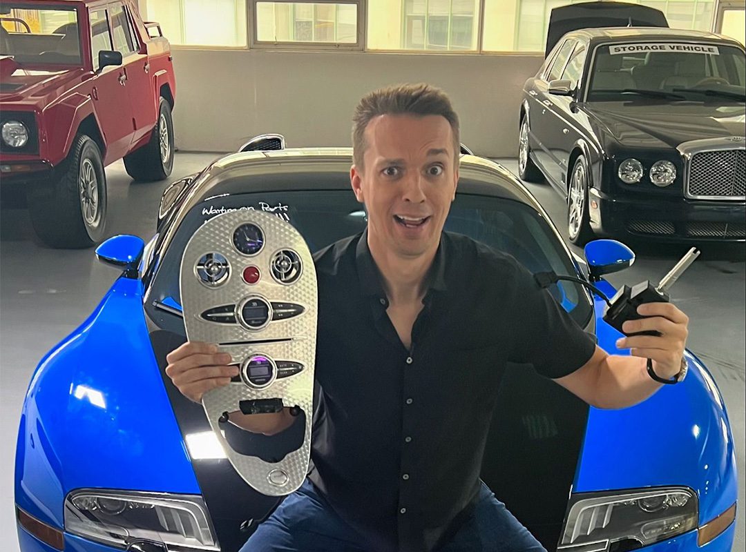 A person holds a car key and panel, sitting on a blue-and-black sports car in a garage filled with luxury cars, including a stunning 2008 Bugatti Veyron from Chicago Motor Cars.
