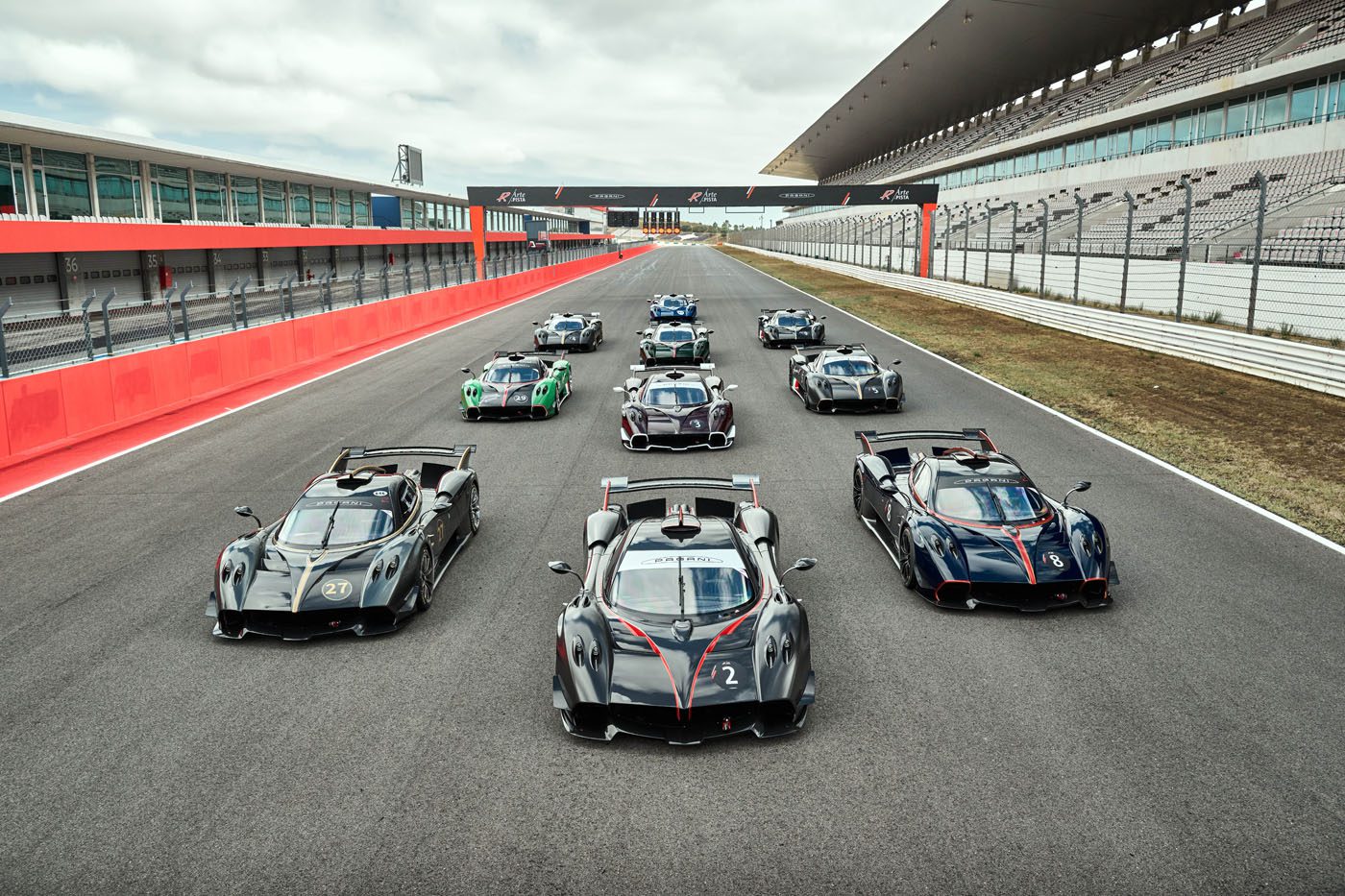 A group of high-performance sports cars is lined up on a race track with grandstands and empty seating visible in the background.