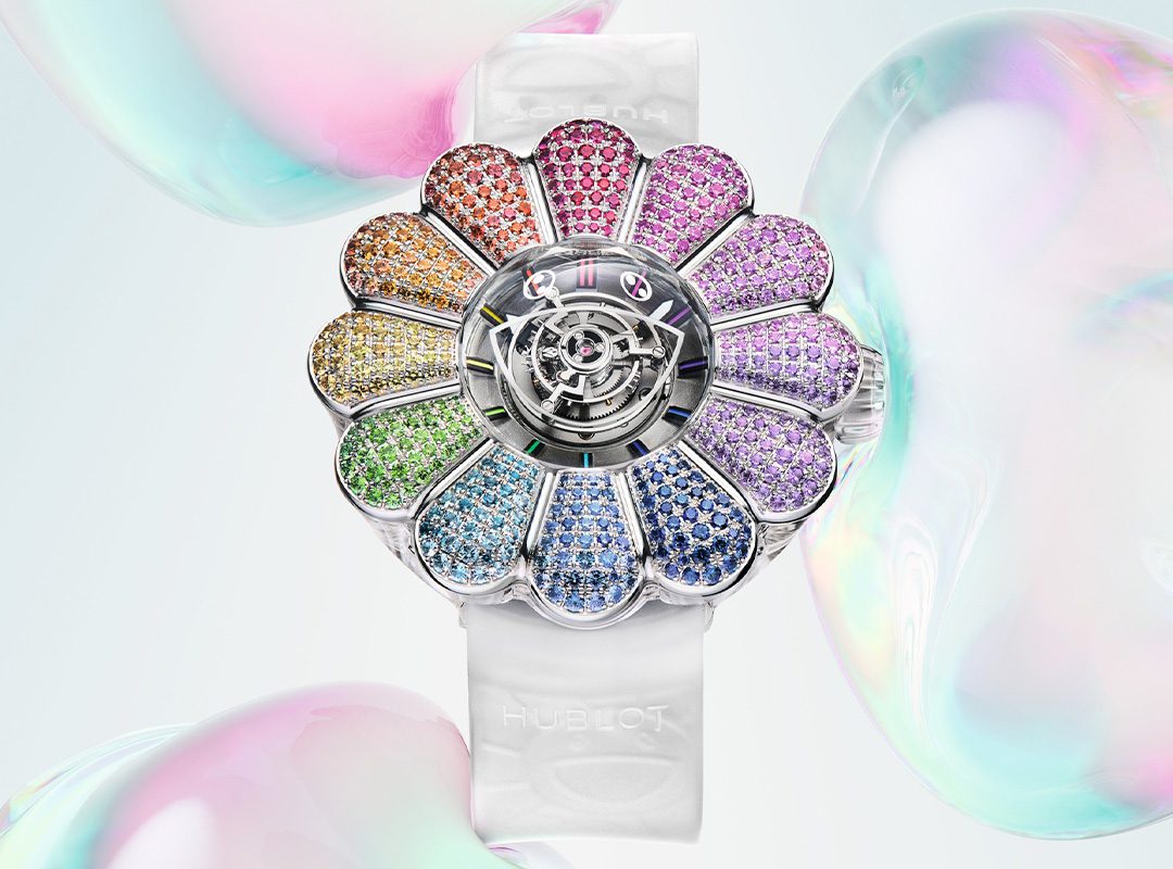 A colorful, Takashi Murakami-inspired flower-shaped jewel-encrusted watch with a white band, set against a soft background with iridescent bubbles.