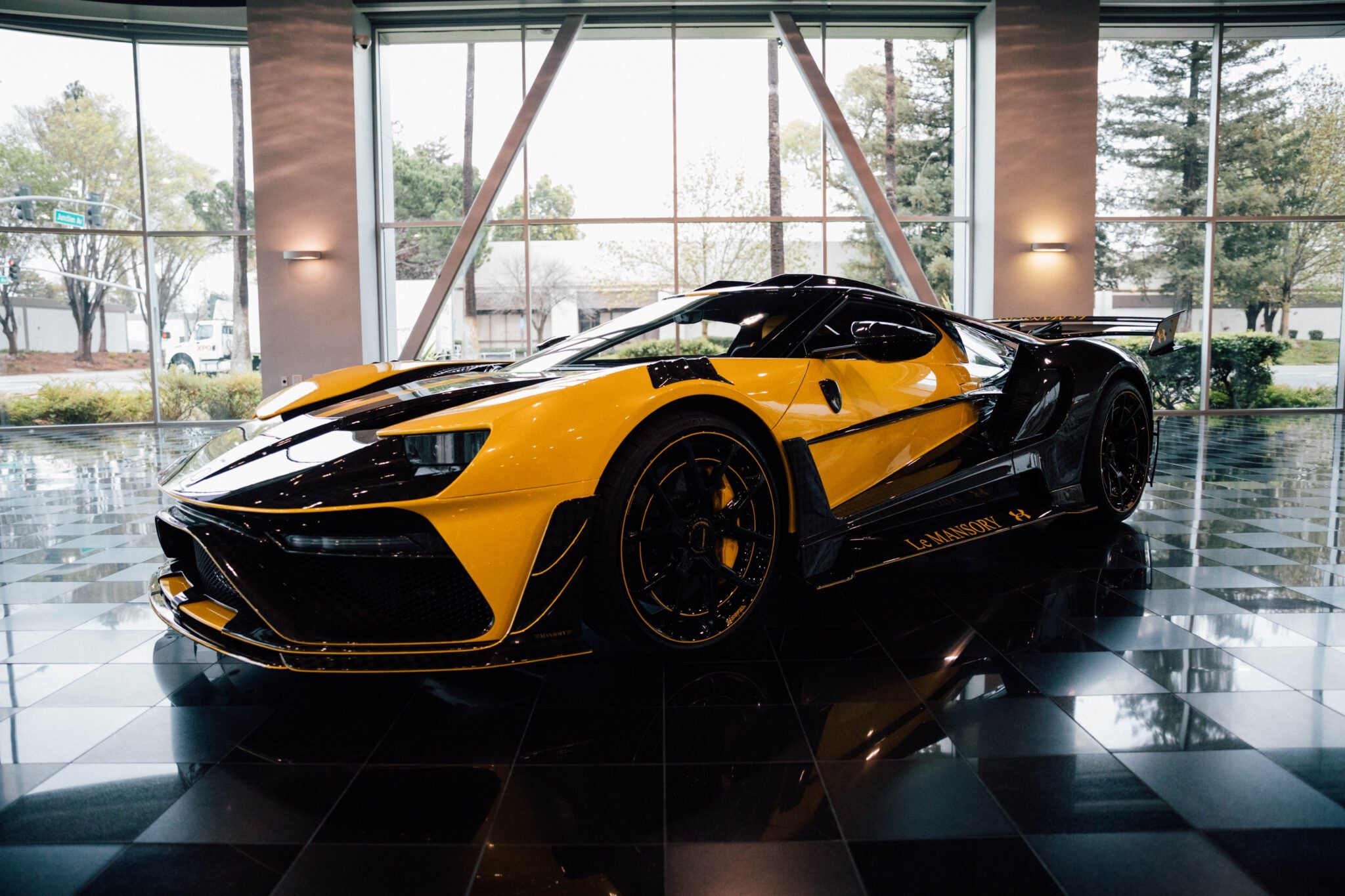 Yellow and black sports car displayed indoors on a reflective black tiled floor, with large windows and trees visible outside.