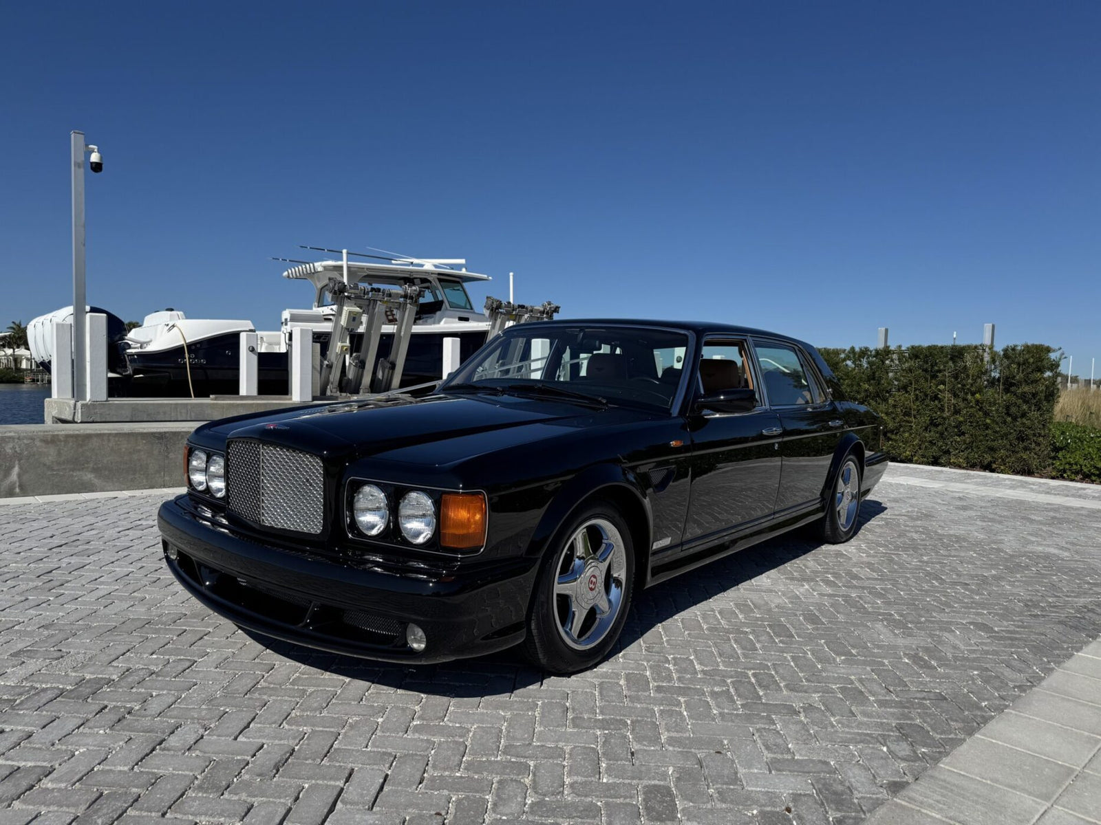 A black Bentley Turbo RT luxury sedan, embodying Mulliner's craftsmanship, sits elegantly on a paved marina with boats in the background under a clear blue sky.