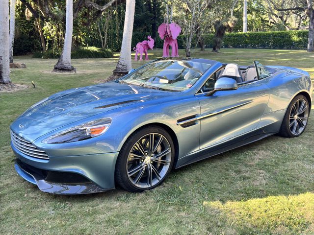 A rare beauty in Ferrari Azzurro Blue, this Aston Martin Vanquish convertible sits elegantly on the grass, with whimsical pink elephant sculptures gracing the background.