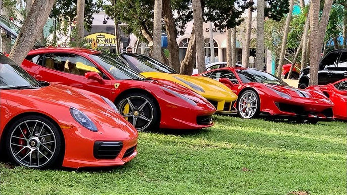 Several red and yellow sports cars, including Ferraris and a Porsche, are parked on grass under trees at the Exotic Car Show in St. Armands Circle, Sarasota.