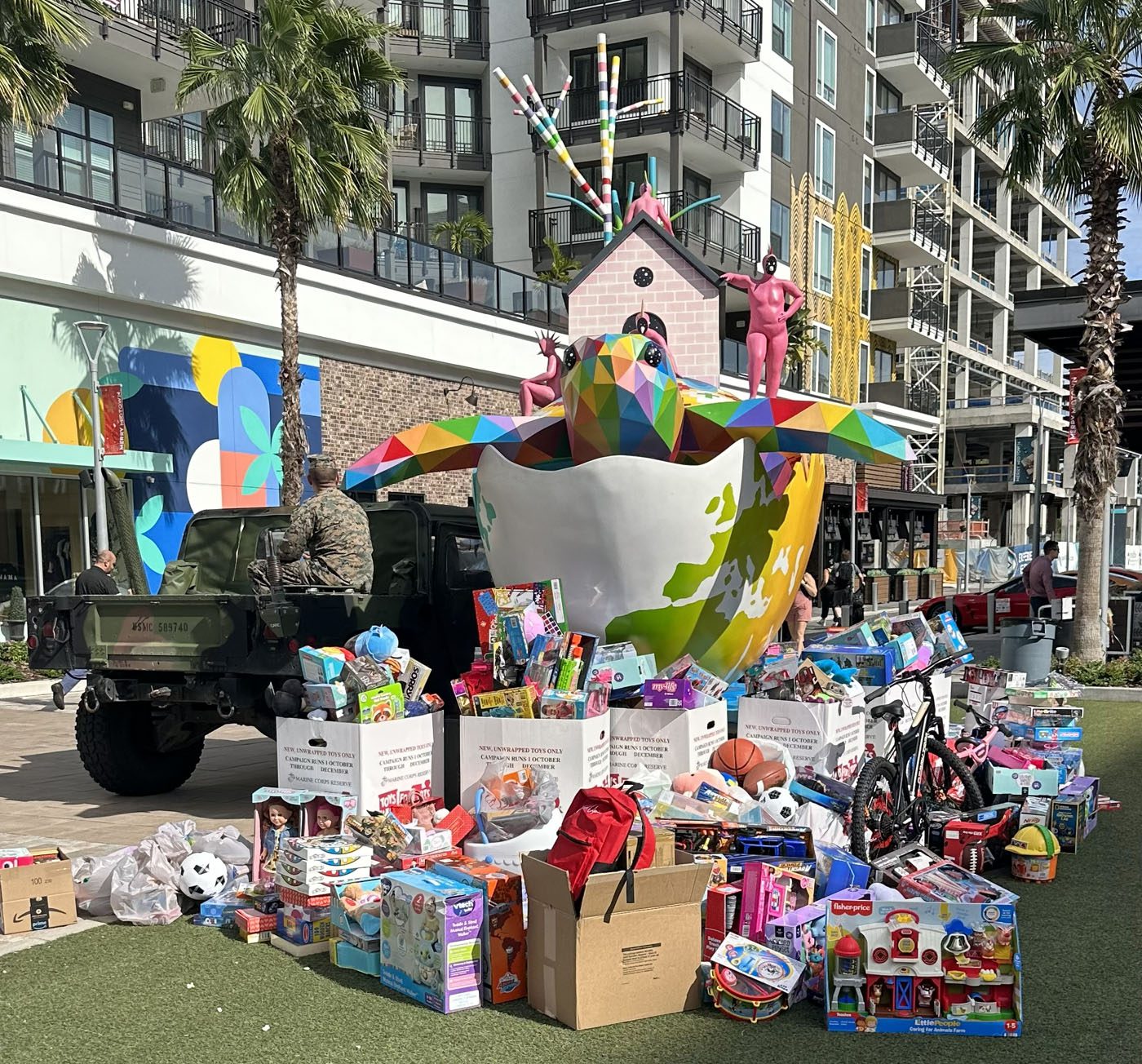 A large, colorful bird sculpture and Toys for Tots donations in boxes and piles are displayed outside a modern apartment building with palm trees.
