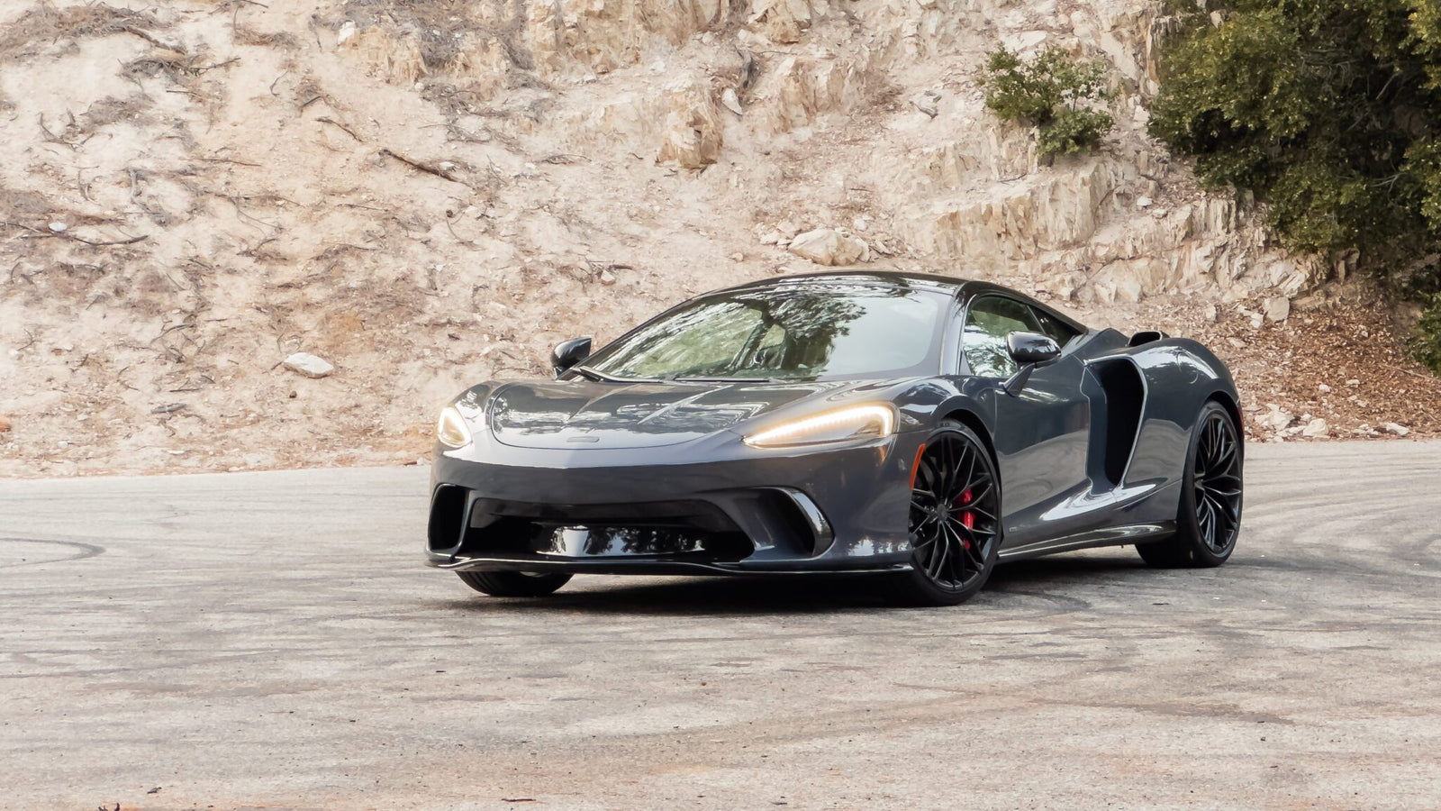 A sleek, gray sports car is parked on a curved road with a rocky, tree-lined backdrop.
