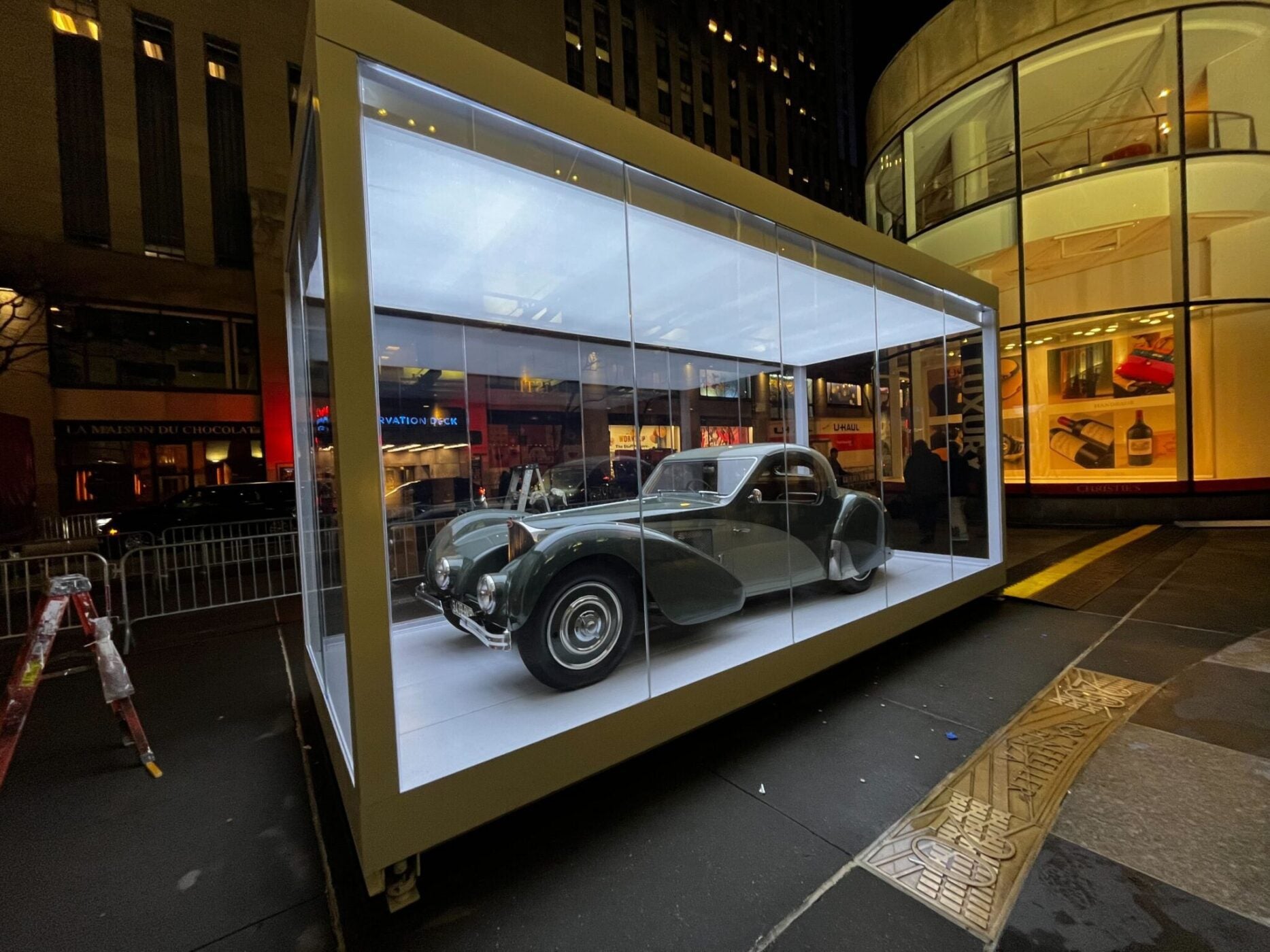 A 1937 Bugatti glistens in a glass case outdoors at night, with Rockefeller Center’s city lights and buildings illuminating the background.