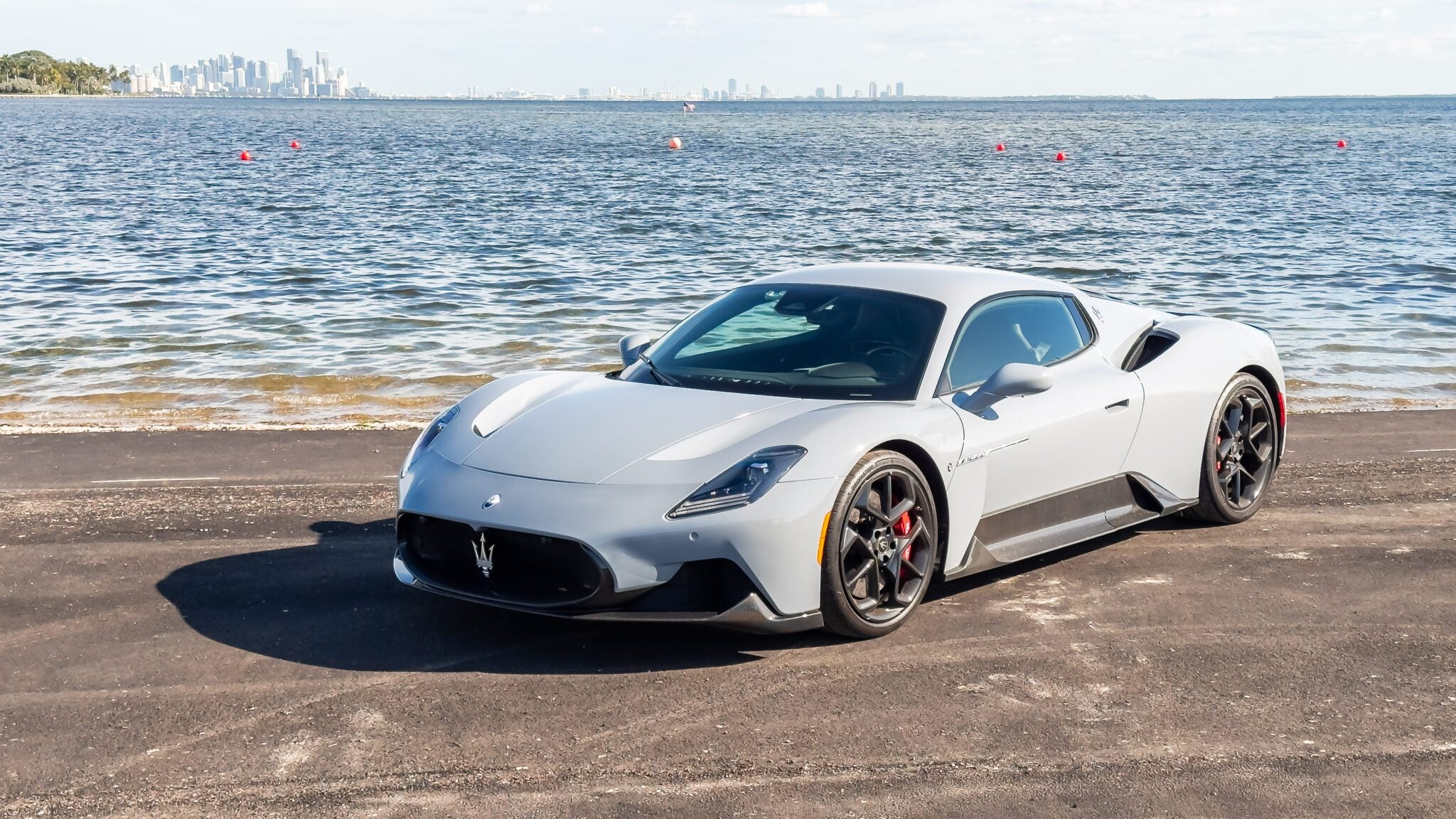 A silver sports car is parked on a paved road by a large body of water with a city skyline in the background.