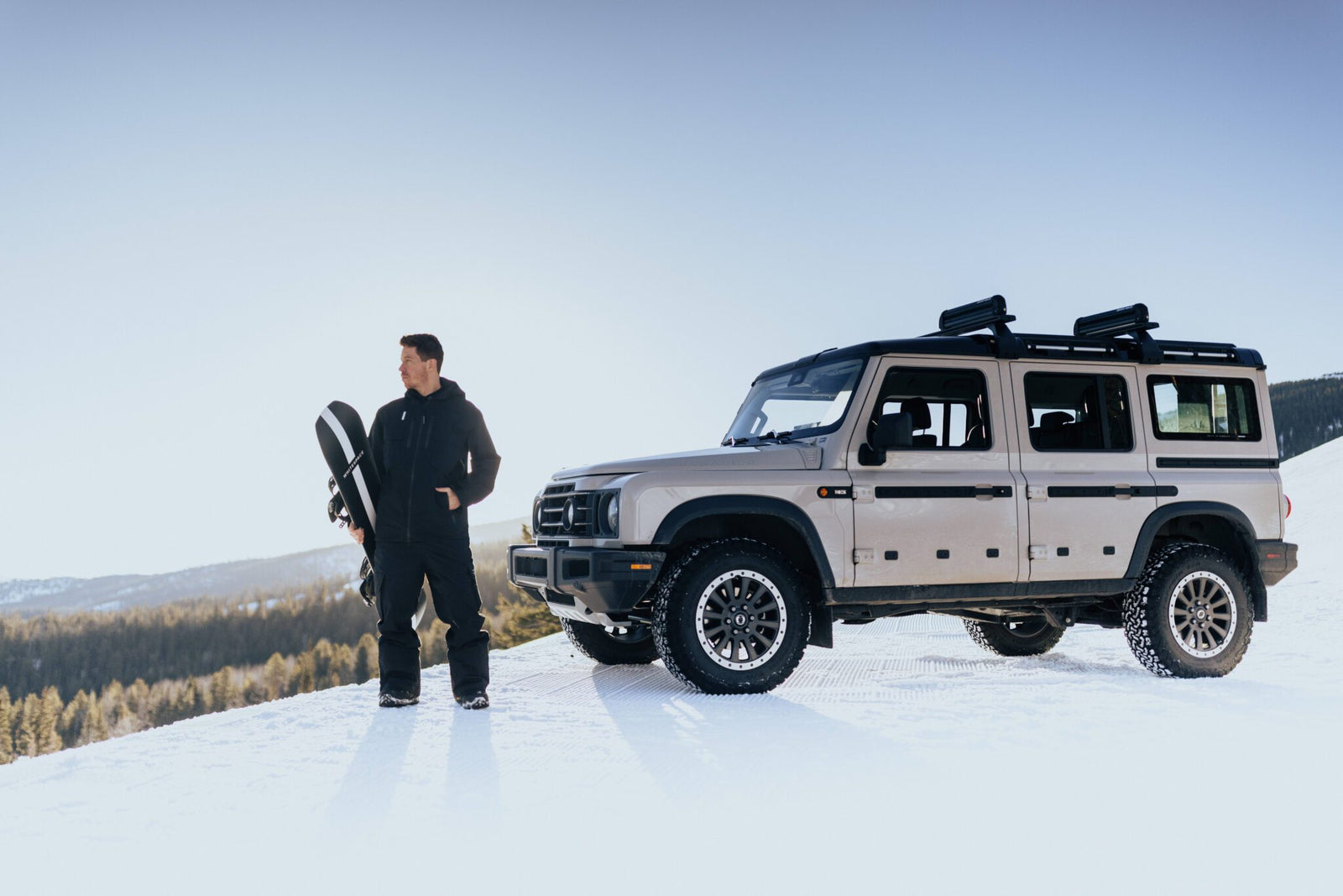 A person stands next to a beige SUV on a snowy hill, inspired by The Snow League, holding a snowboard under a clear blue sky.