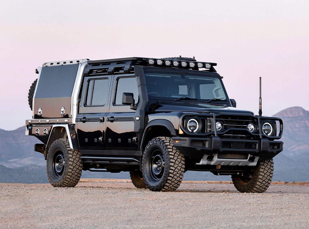 The INEOS Grenadier Quartermaster is a rugged black off-road vehicle with a custom build, featuring large tires and additional lighting, parked on a dirt terrain.