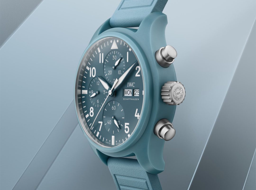 A blue IWC Miramar Pilot’s Chronograph wristwatch with a blue rubber strap, showing the time 10:08, date 25, and day Monday, set against a matching blue geometric background.