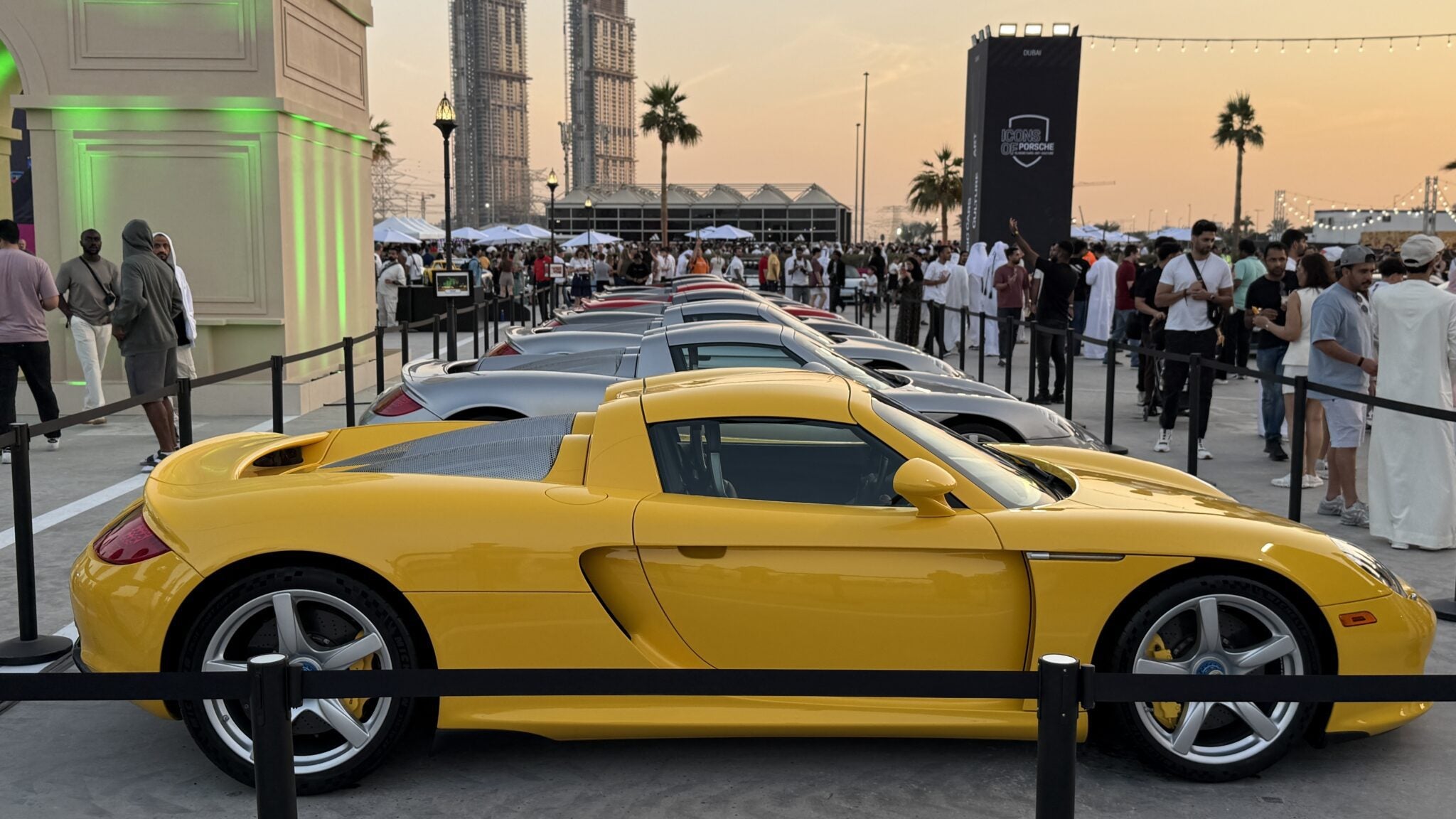 Feature Exclusive: 2025 Icons of Porsche Dubai—a row of sports cars, led by a yellow Porsche Carrera GT, is displayed at an outdoor event attended by a crowd of people.
