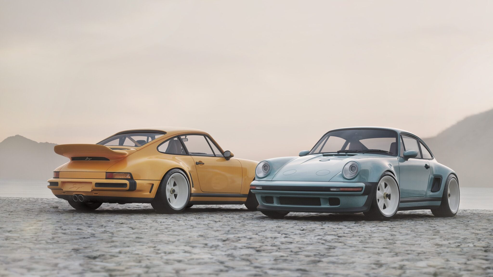 Two vintage sports cars, including a limited edition Porsche 911 by Singer in yellow and a classic blue model, are parked side by side on a cobblestone surface near a misty lake with mountains in the background.