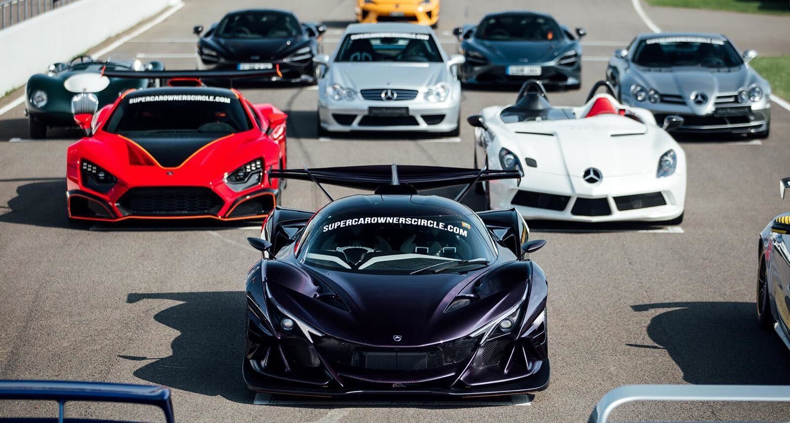 A lineup of high-performance supercars, including a black hypercar in front, parked on a racetrack under clear daylight.