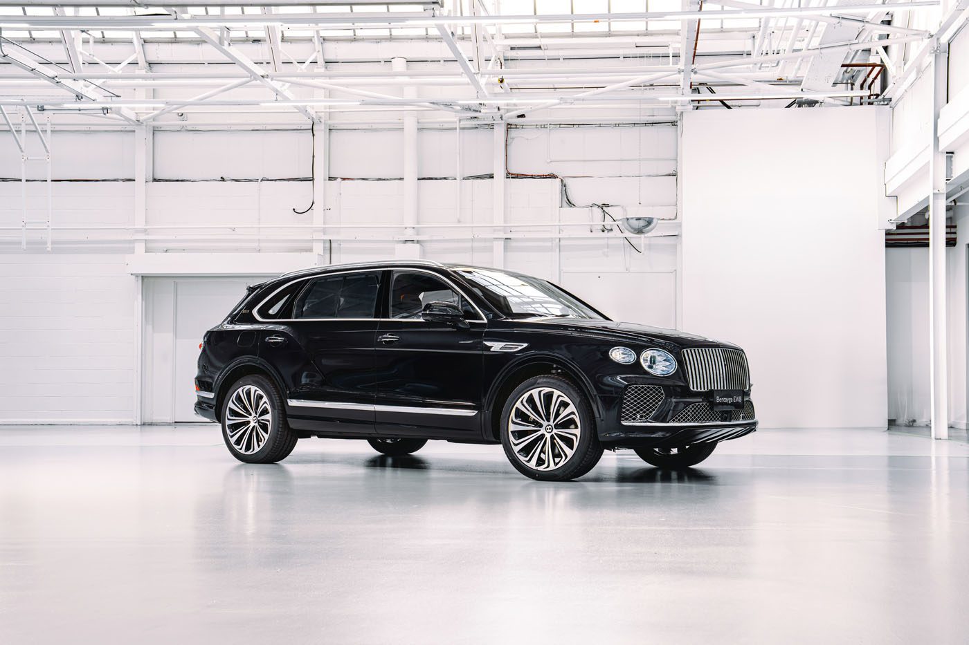A black Bentley luxury SUV, part of the Mulliner Bespoke Collections, is parked in a spacious, white industrial-looking room with visible ceiling beams.