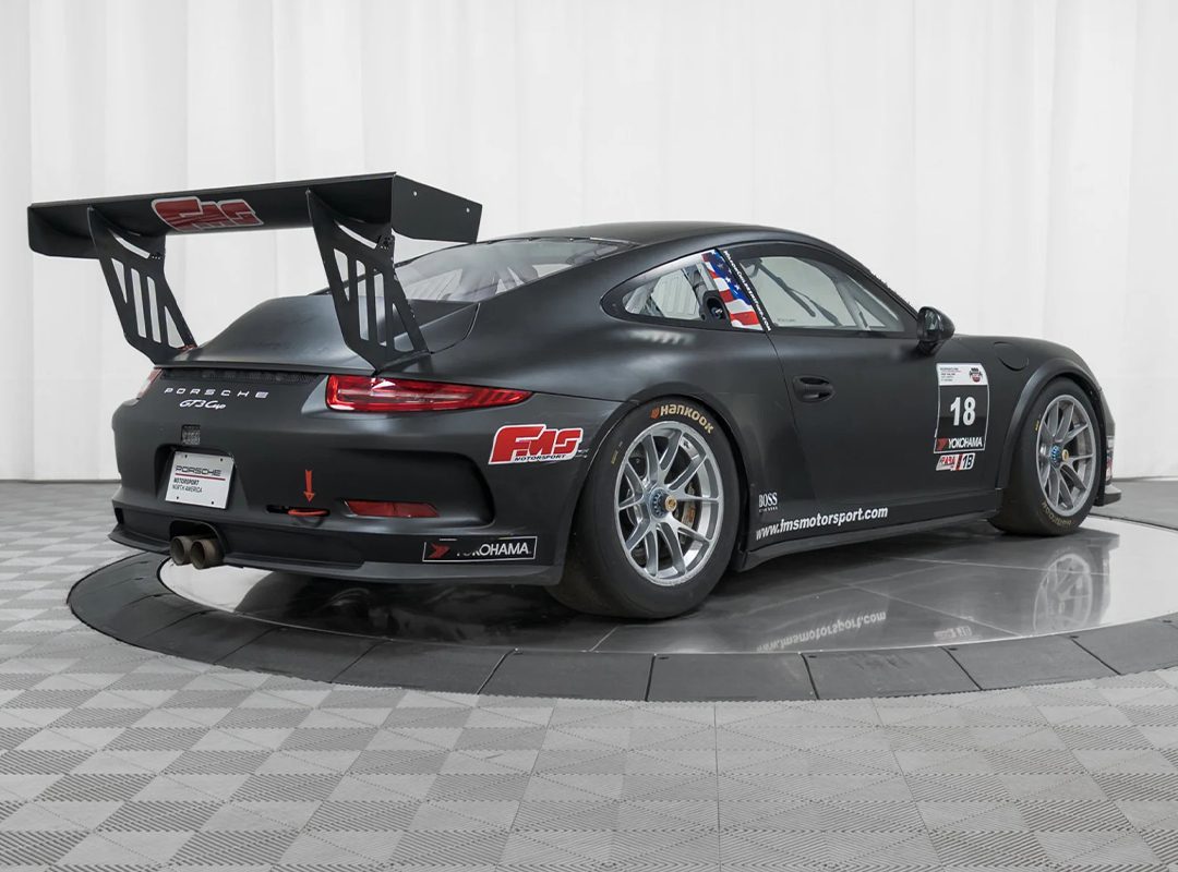 A matte black Porsche 911 GT3 race-ready car with a large rear wing, racing decals, and number 18, displayed in a studio with a checkered floor and white background. Offered by Isringhausen Imports for true Porsche track car enthusiasts.