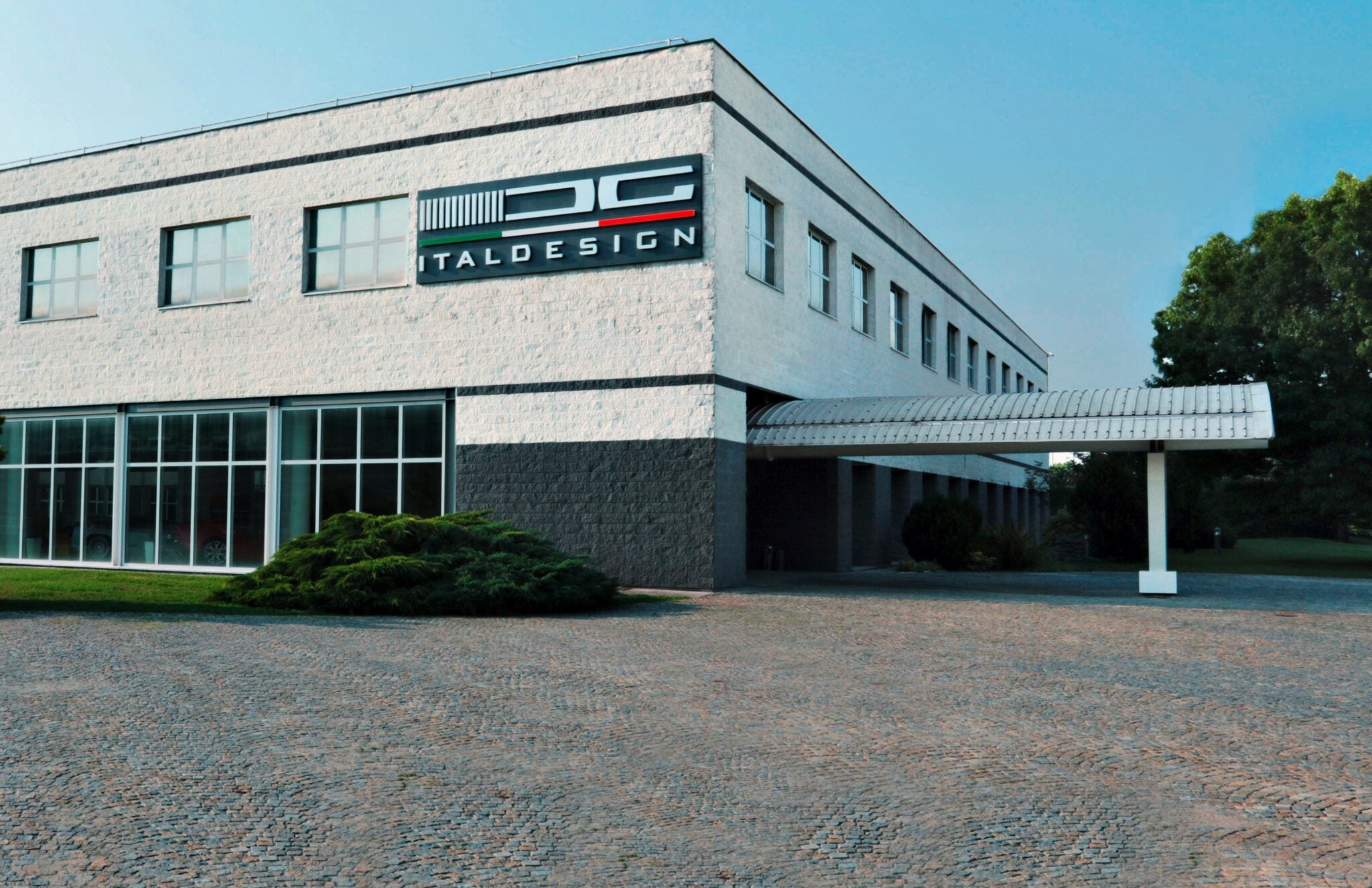 A modern two-story building with large windows and "Italdesign" signage on the upper corner facade, surrounded by greenery and a cobblestone driveway, now highlights California-based UST takes majority control of Italdesign.