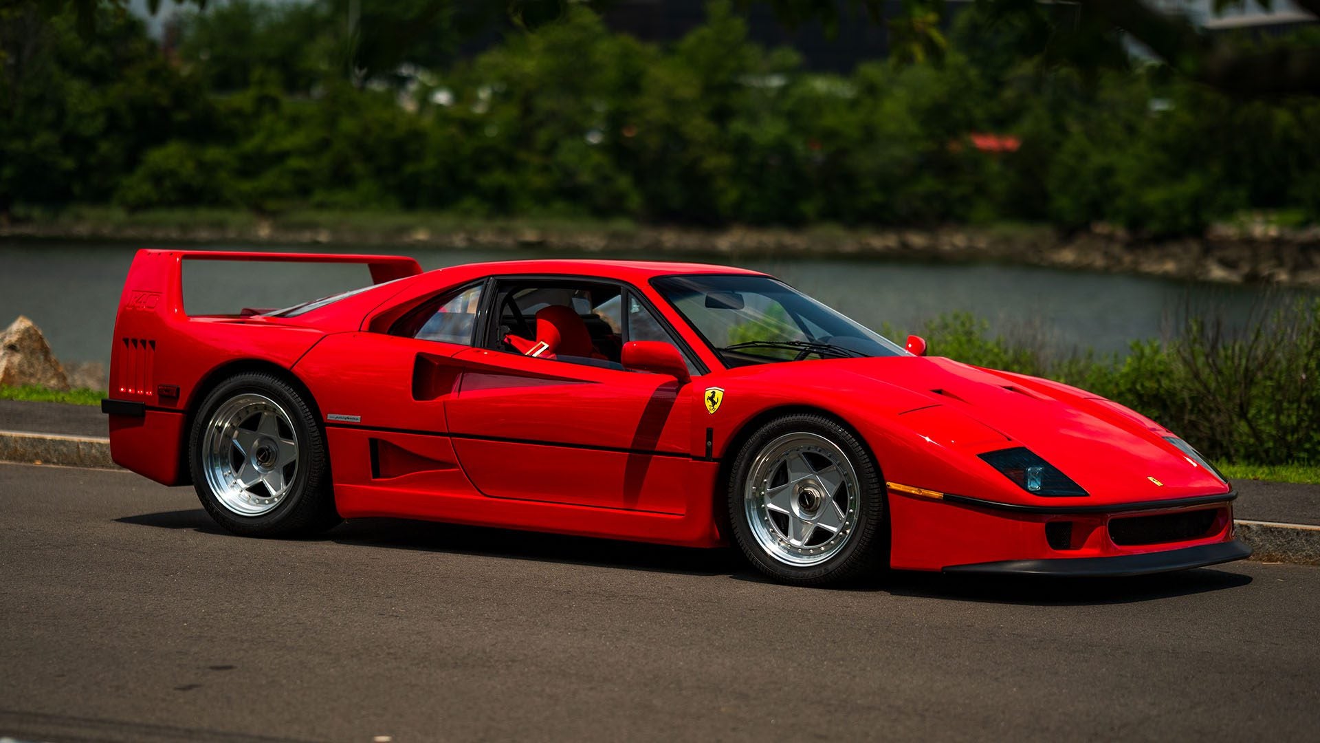 A 1991 Ferrari F40 sports car is parked on a paved road with greenery and a body of water in the background, reminiscent of those showcased at the Monterey Jet Center Auction.