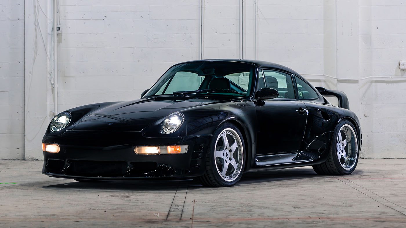 A black 1998 RUF CTR 2 Sport with a wide body kit and custom wheels is parked indoors against a white brick wall, ready to turn heads at the Monterey Jet Center Auction.