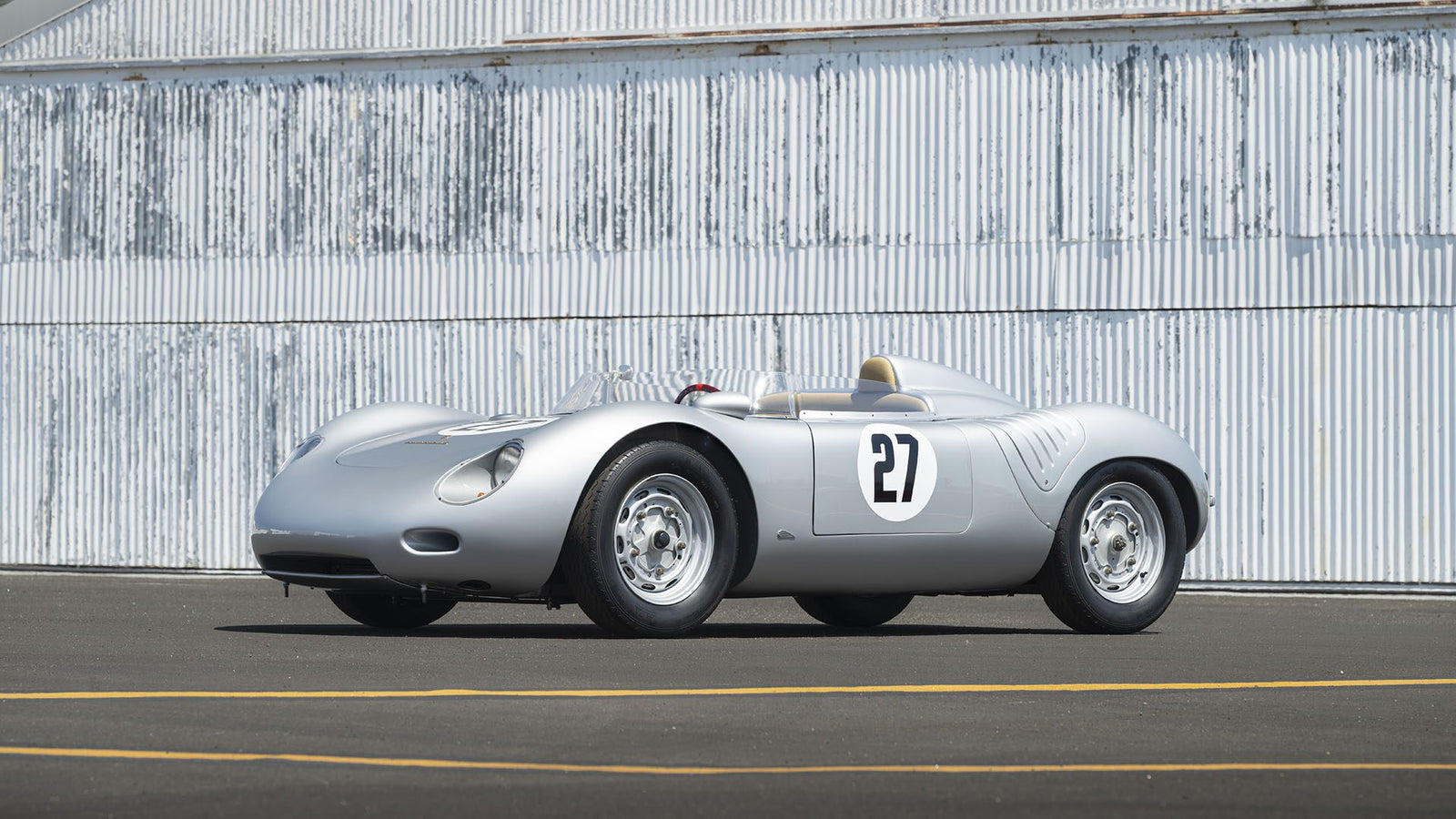 A silver Porsche 718 RSK Spyder vintage race car with the number 27 on its side is parked on an asphalt surface in front of a corrugated metal wall at the Monterey Jet Center Auction 2025.