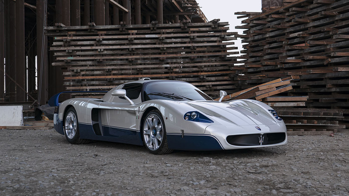 A striking white and blue 2005 Maserati MC12 sports car is parked on a gravel surface between large stacks of wooden planks and industrial beams at the Monterey Jet Center Auction.