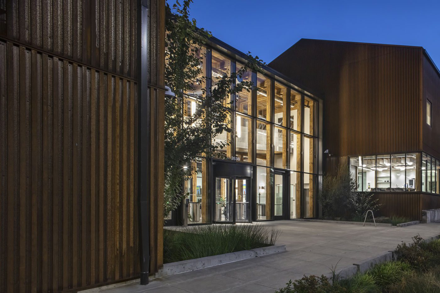 Modern building with large glass windows and wooden panels, surrounded by greenery, under a clear evening sky.