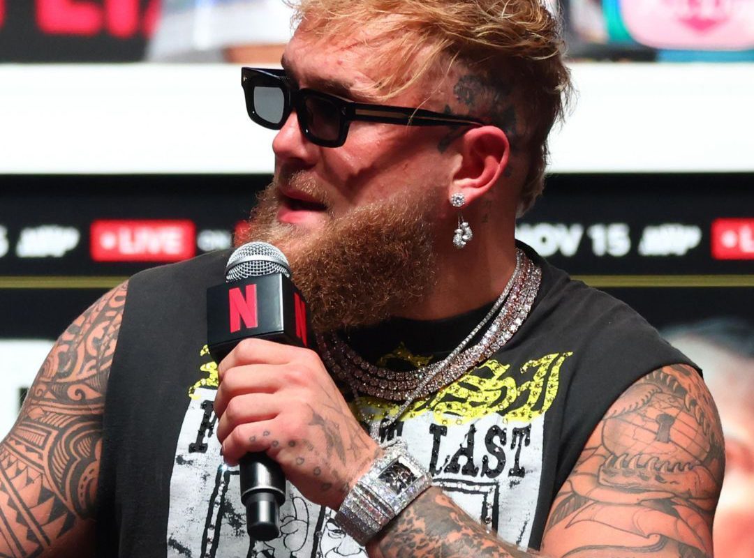 A man with sunglasses and tattoos holds a microphone, wearing a printed sleeveless shirt, multiple necklaces, and earrings. He sports a $7M watch while channeling Jake Paul's bold style.