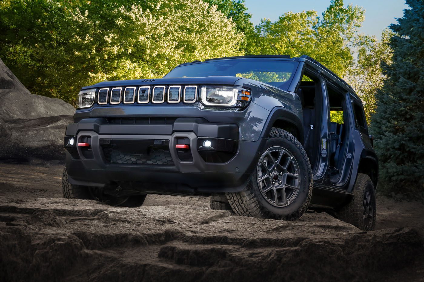 A trail-ready 2026 Jeep Recon, all-electric and dark gray, with no doors or roof panels, is parked on rocky terrain surrounded by trees and greenery under a clear sky.