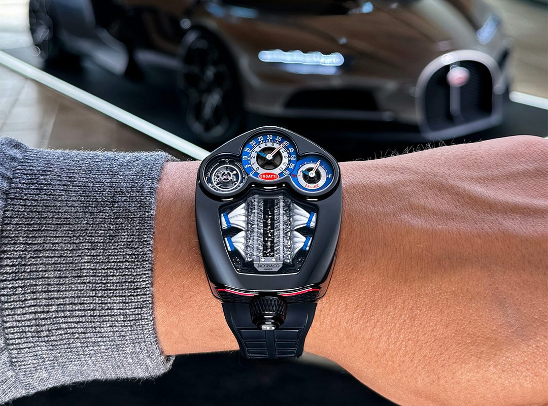 A close-up of a person’s wrist wearing a futuristic Jacob & Co. Bugatti Tourbillon watch, inspired by automotive design, with a luxury sports car blurred in the background.