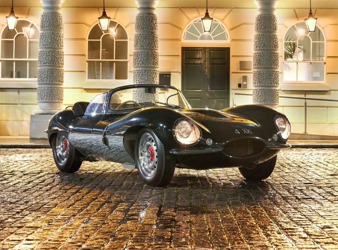A vintage 1957 Jaguar XKSS sports car is parked on a wet cobblestone street, with an elegant building in the background, its allure capturing the essence of classic luxury—an icon that once fetched $14 million at an RM Sotheby's Auction.