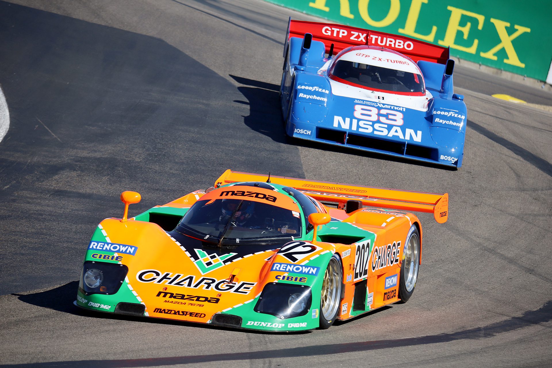 Two vintage race cars—a green-orange Mazda and a blue Nissan—celebrate Japanese Motorsports Heritage as they speed past a green Rolex ad at the 2026 Rolex Monterey Motorsports Reunion.