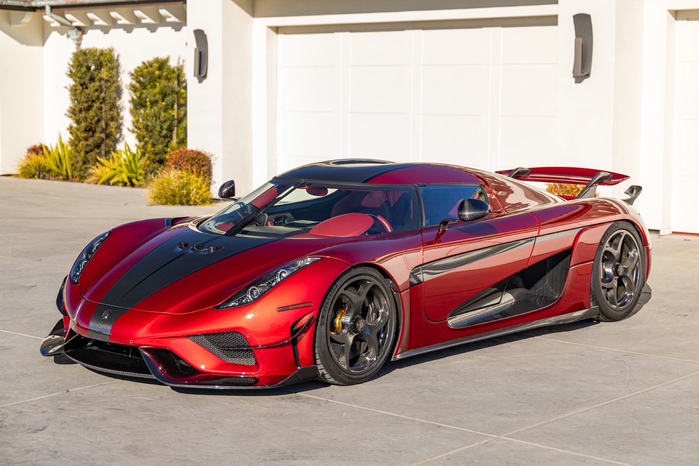 A Koenigsegg Regera is parked outside a modern building with white garage doors and lush plants nearby, showcasing its extreme car design in striking red and black.