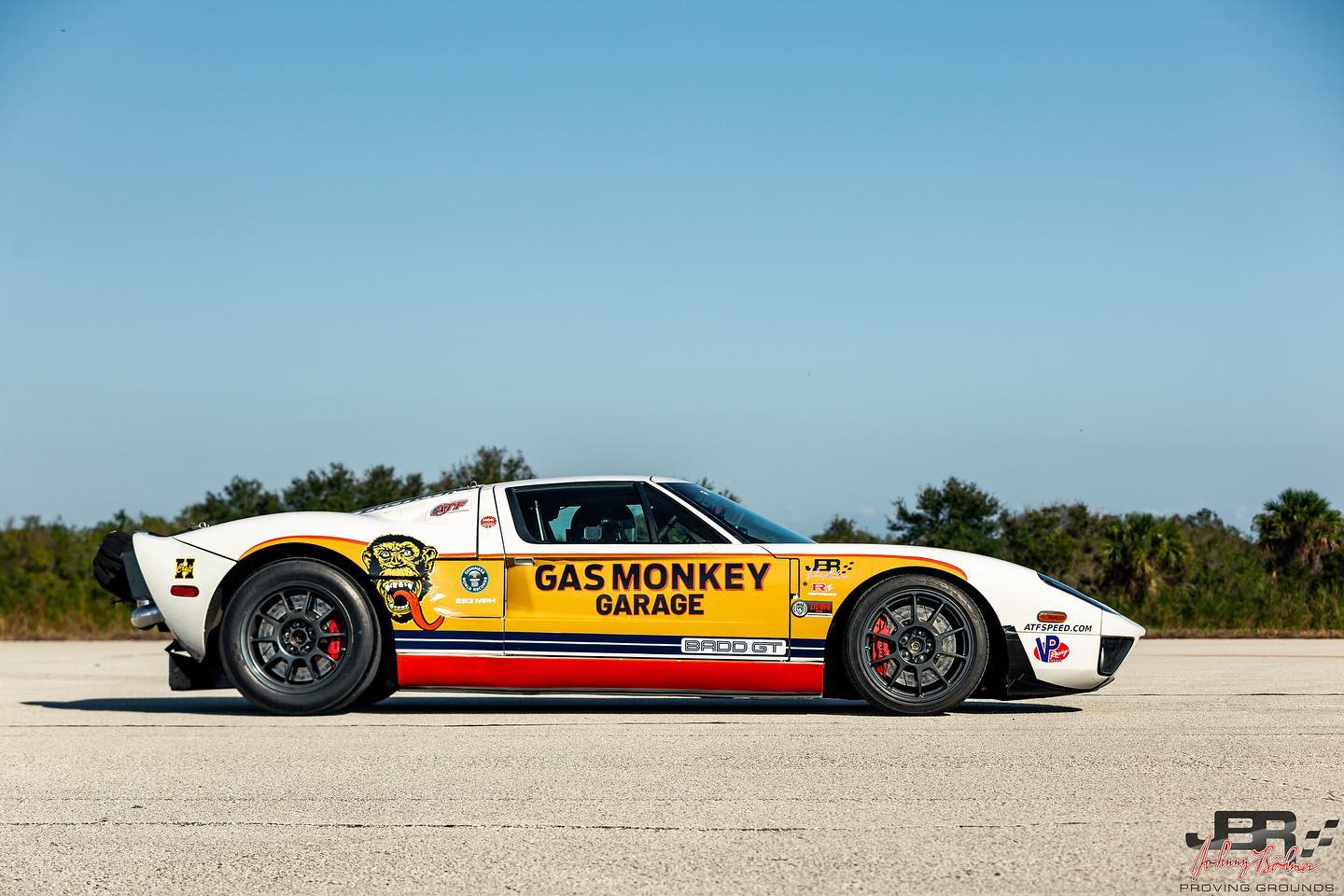 A yellow and white Ford GT sports car, known as the BADD GT and driven by Jonny Bohmer, with "Gas Monkey Garage" branding, is parked on a paved surface under a clear sky.