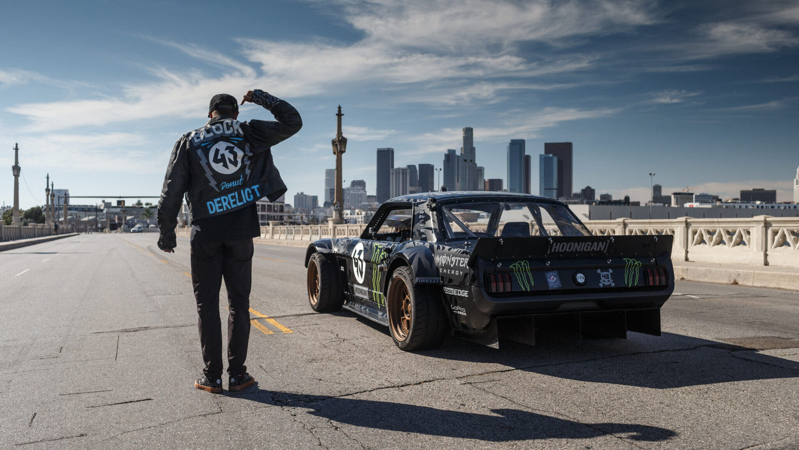 A person stands next to a modified black race car on an empty street with a city skyline in the background.