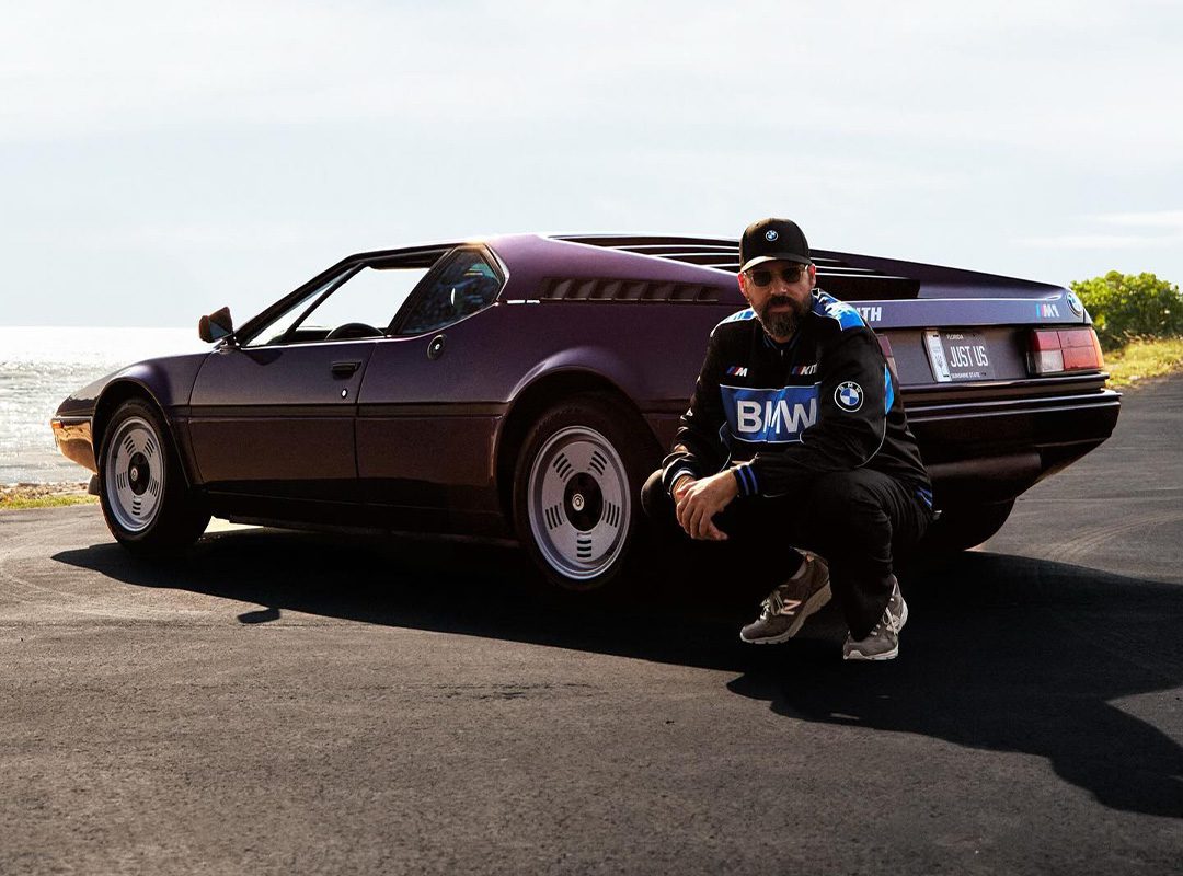 A man crouches beside a vintage purple sports car, reminiscent of Ronnie Fieg's collaborations, with custom license plate "MLS 16" on a sunny road by the sea.