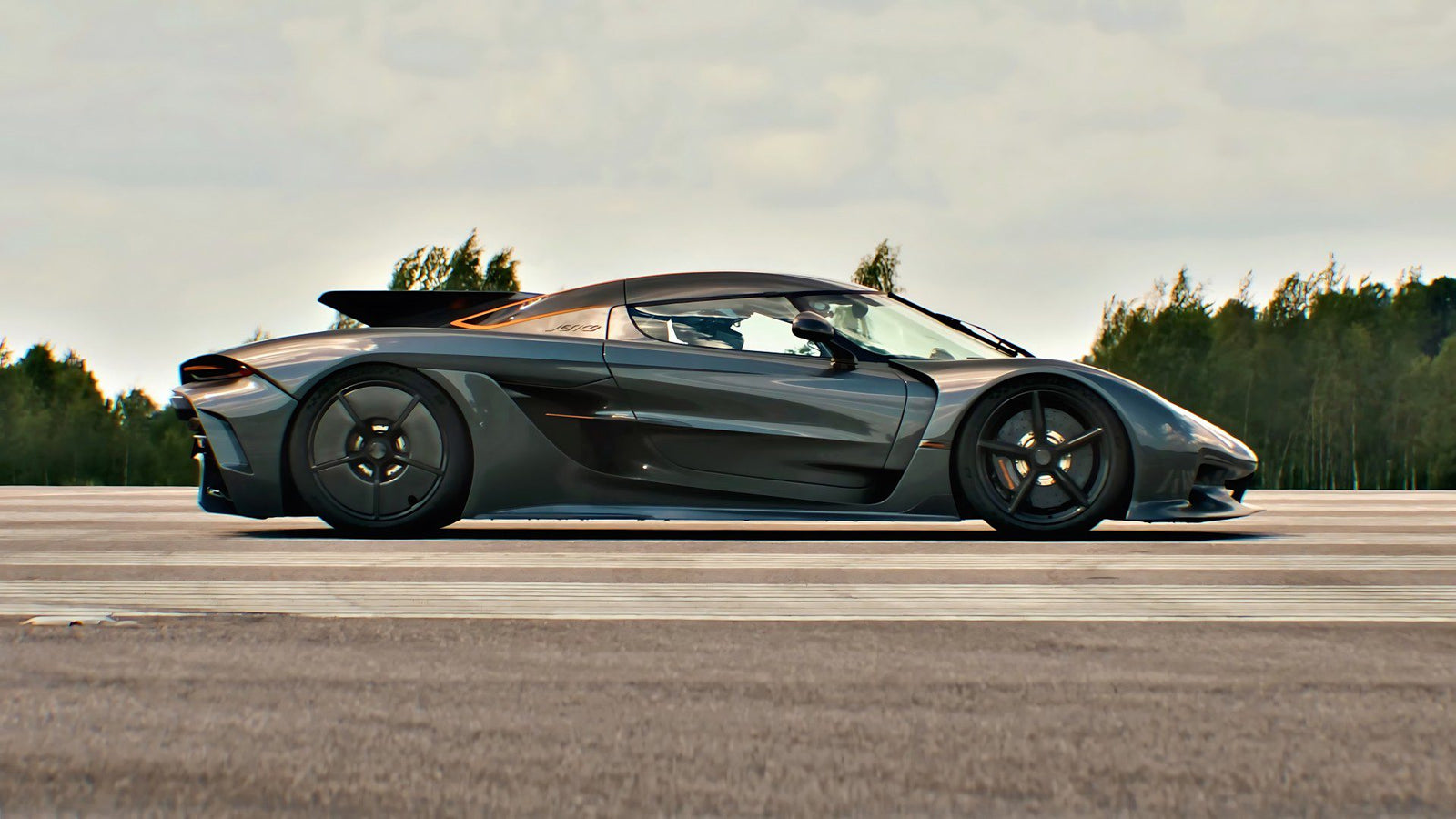 A sleek black sports car, reminiscent of the Koenigsegg Jesko Absolut, is parked on a road lined with trees under a cloudy sky, exuding power worthy of the 1/2 Mile Top Speed Record.
