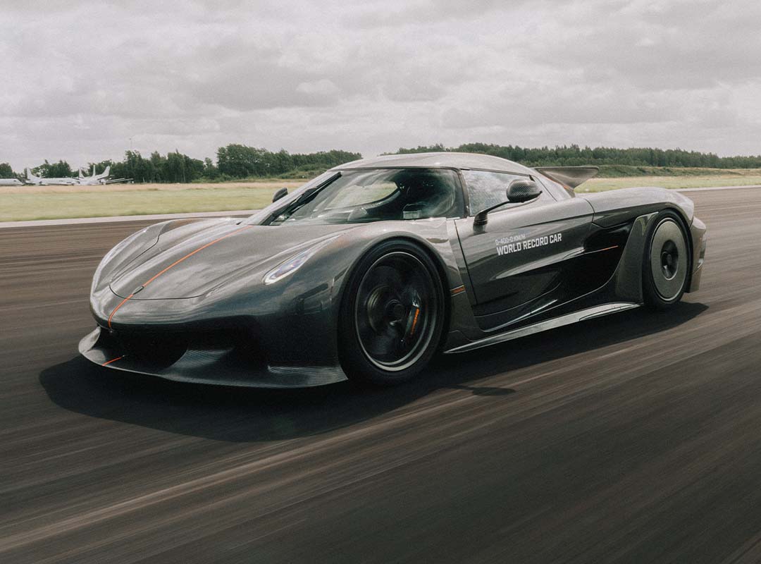 A grey Koenigsegg Jesko Absolut with "World Record Car" text on the door speeds along a racetrack, set against a grassy landscape and cloudy sky, reflecting its 0-250-0 MPH world record capabilities.