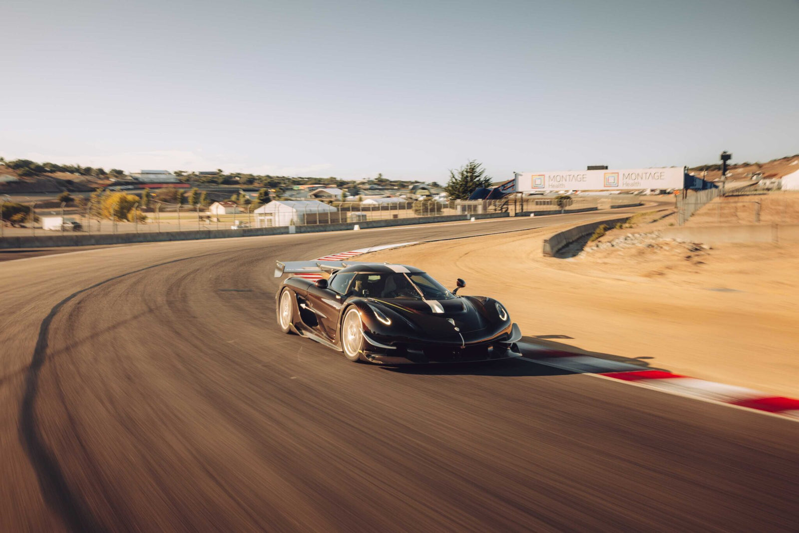 A black Koenigsegg Sadair’s Spear speeds around a curve on a sunny racetrack, shattering the Laguna Seca production car record for 2025, with barriers, signage, and hills visible in the background.