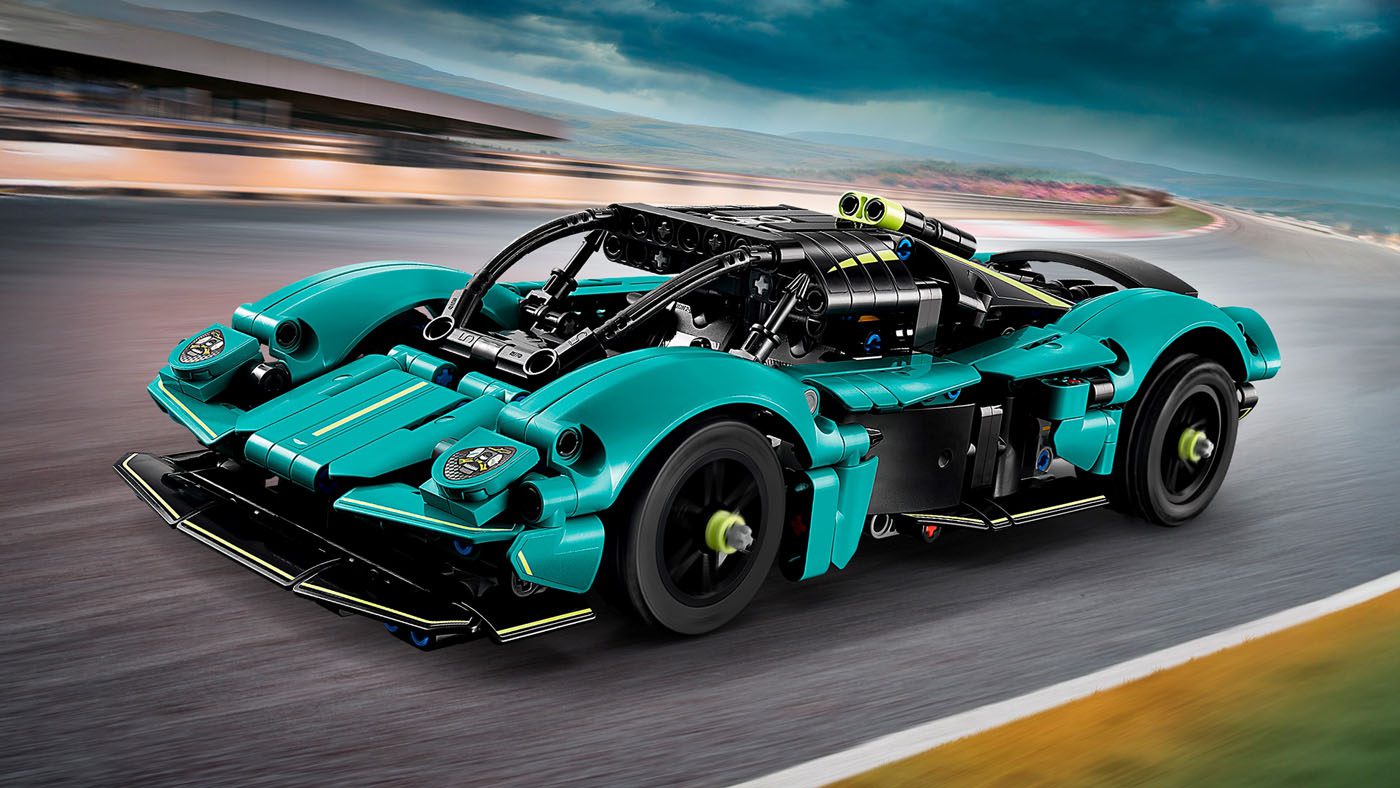 A teal and black LEGO Technic toy race car model with detailed mechanical features, inspired by the Aston Martin Valkyrie, drives on a blurred racetrack under a cloudy sky—perfect for fans looking to buy an exciting collectible.