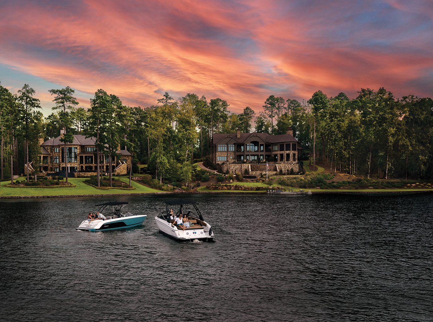 Two boats glide across a lake near upscale lakeside houses in the renowned Reynolds Lake Oconee community, surrounded by lush trees at sunset. The vibrant, orange-pink sky above adds to the charm of what many consider the South's Best Lake.