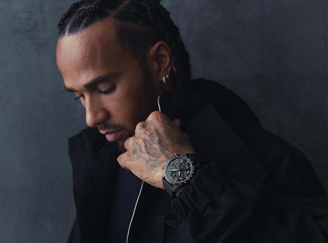Person with braided hair and tattoos looks down, wearing a dark jacket and an IWC Watch, reminiscent of Lewis Hamilton's style.