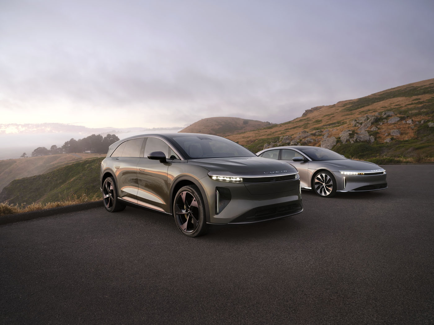 Two sleek, modern cars, including the 2025 Lucid Gravity, parked majestically on a scenic road with hills and a cloudy sky overhead.