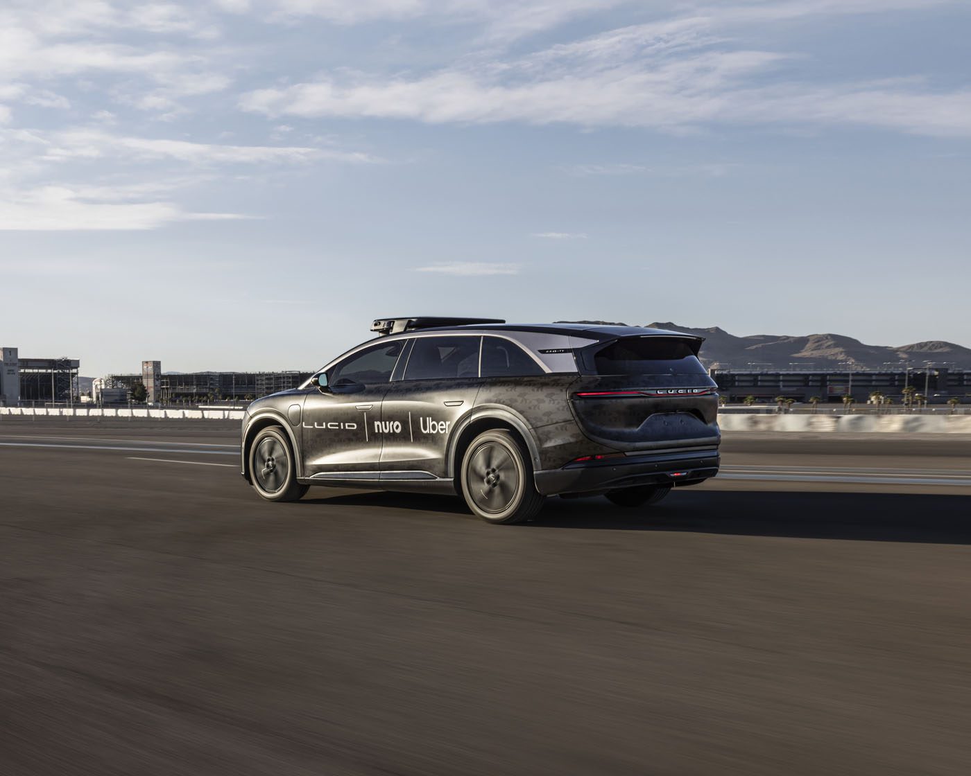 A black SUV with logos including Lucid, Nuro, and Uber drives on a highway, with city buildings and mountains in the background.