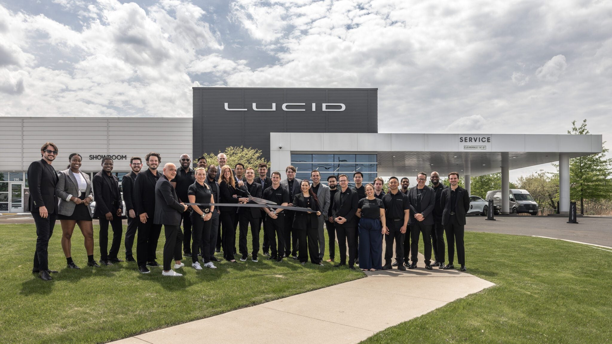 A group of people poses in front of a Lucid building and service center, with several individuals holding a large ceremonial ribbon-cutting scissors.