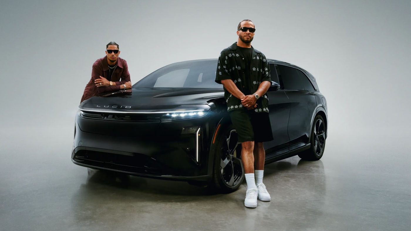 Jalen Brunson and Josh Hart, both wearing sunglasses, stand in front of a black Lucid electric vehicle in a studio setting for a Lucid campaign.