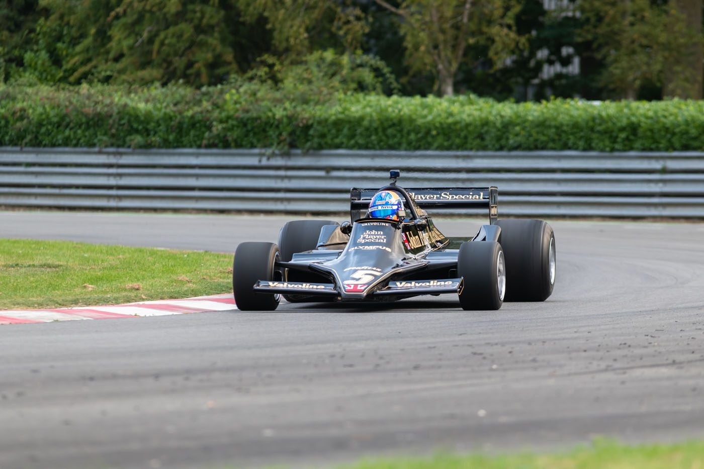 A black Lotus Type 79 Formula 1 car with John Player Special livery takes a corner on a racetrack, driven by Jan Lammers wearing a blue helmet.