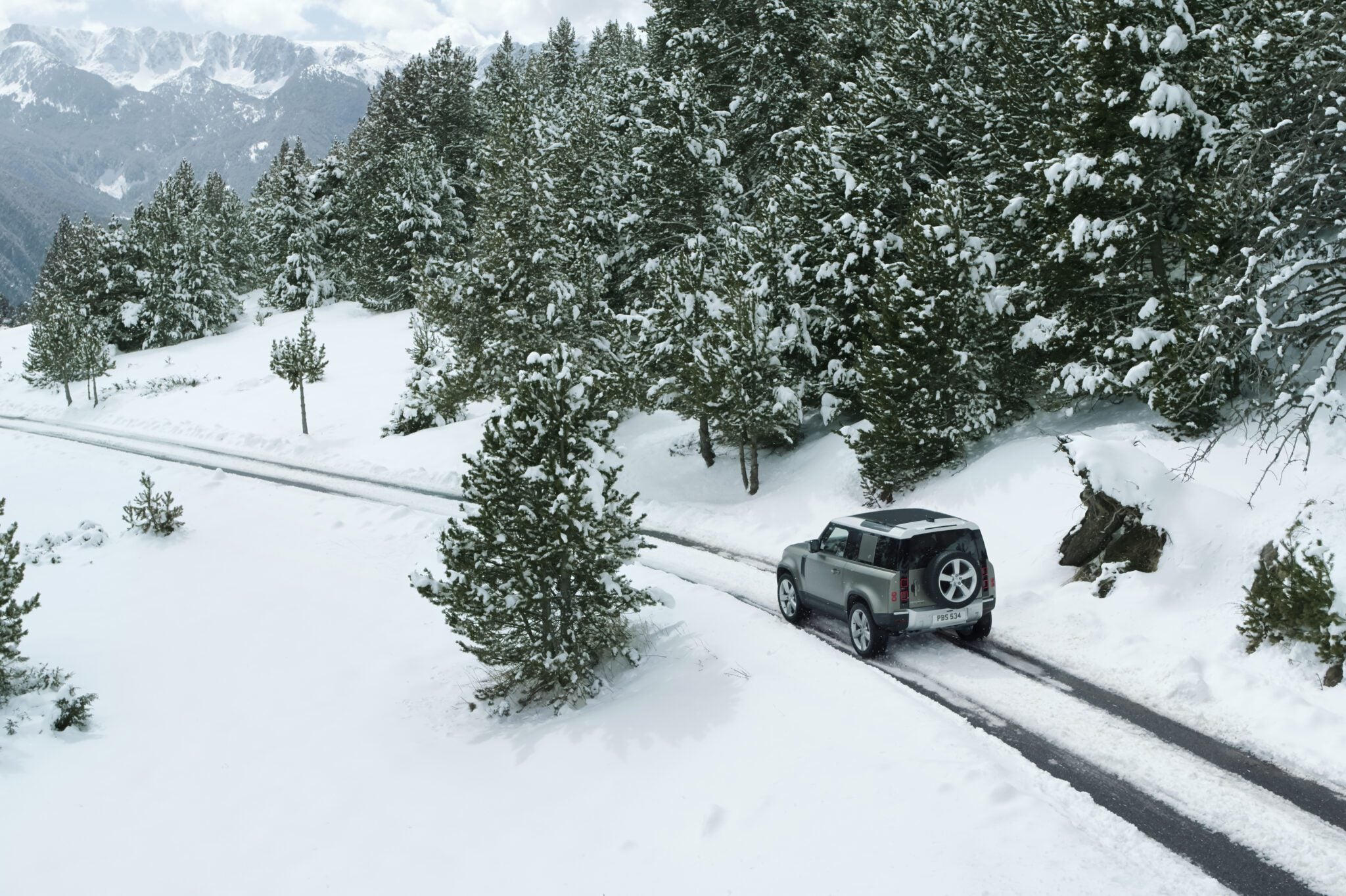 A silver SUV navigates a narrow, snow-covered road amid dense pine trees and majestic mountains, braving snowstorm conditions with determination.