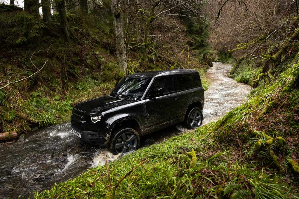 A black SUV drives through a shallow stream surrounded by green grass and trees in a forested area during the Destination Defender Festival in New York’s scenic Hudson Valley.